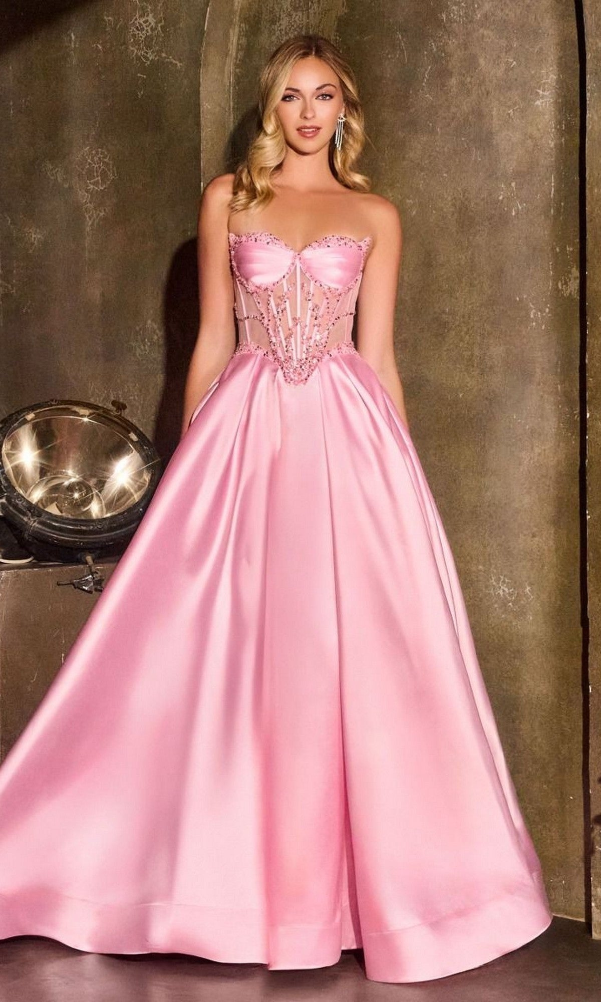 A woman stands against a textured wall, wearing the Ladivine CD389—a floor-length pink strapless Mikado prom dress with an A-line skirt, corset-style bodice, and sparkling embellishments.